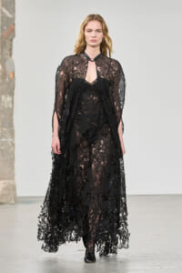 Runway model wearing a full-length black lace gown with sheer overlay and long sleeves, mid-stance on a fashion show runway, neutral expression.