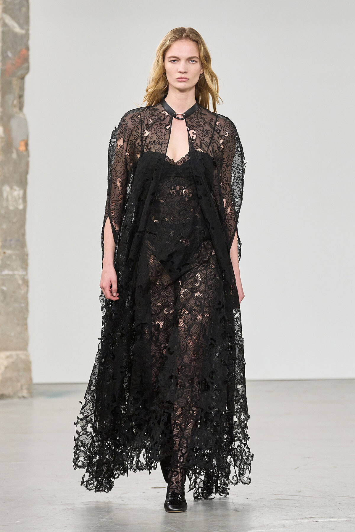 Runway model wearing a full-length black lace gown with sheer overlay and long sleeves, mid-stance on a fashion show runway, neutral expression.