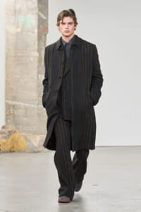 Male model walking a fashion show in a dark pinstripe overcoat and matching suit, hands in pockets, neutral expression on a minimal runway.