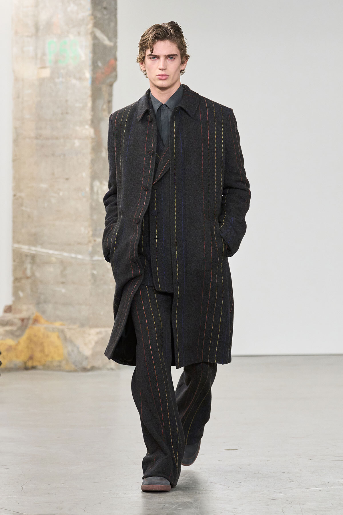 Male model walking a fashion show in a dark pinstripe overcoat and matching suit, hands in pockets, neutral expression on a minimal runway.