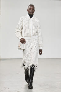 Model walking runway in a white crochet-knit jacket and matching fringed skirt with a white bag and black boots.