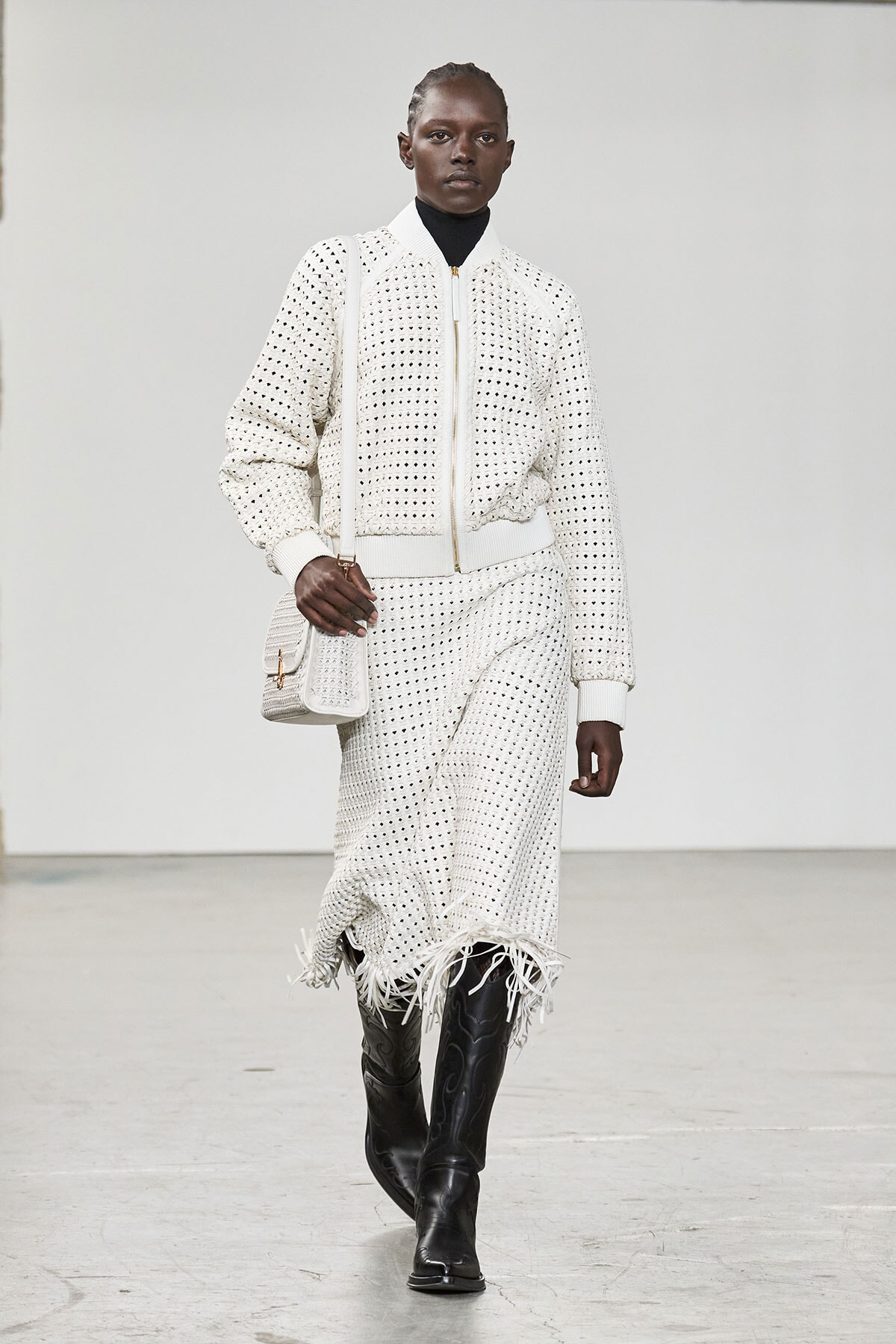 Model walking runway in a white crochet-knit jacket and matching fringed skirt with a white bag and black boots.