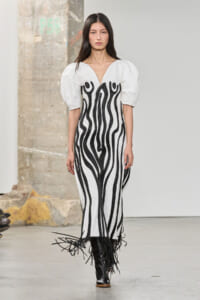 Model walks a runway in a white gown with black wavy embroidery and fringe at the hem, paired with black boots.