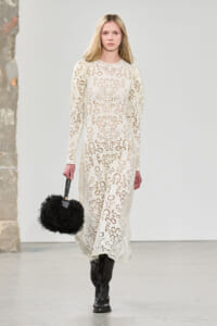 Runway model in a cream lace dress, carrying a black fluffy handbag and wearing black boots on a minimalist set.