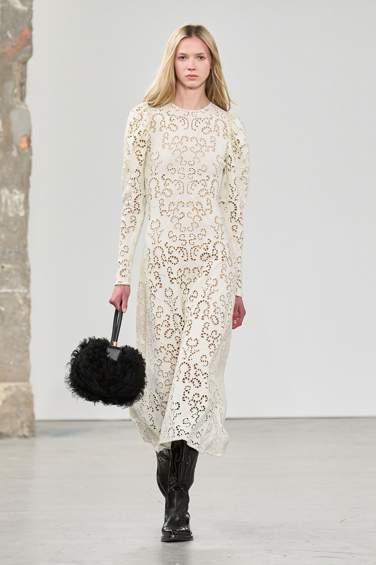 Runway model in a cream lace dress, carrying a black fluffy handbag and wearing black boots on a minimalist set.