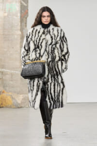 Model walking wearing a white and black zebra-pattern fur coat, black turtleneck, knee-high black boots, and a black crocodile-textured handbag with gold hardware.
