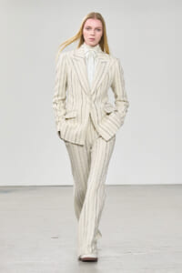 Model wearing a cream pinstripe suit (blazer and wide-leg trousers) walking on a runway.