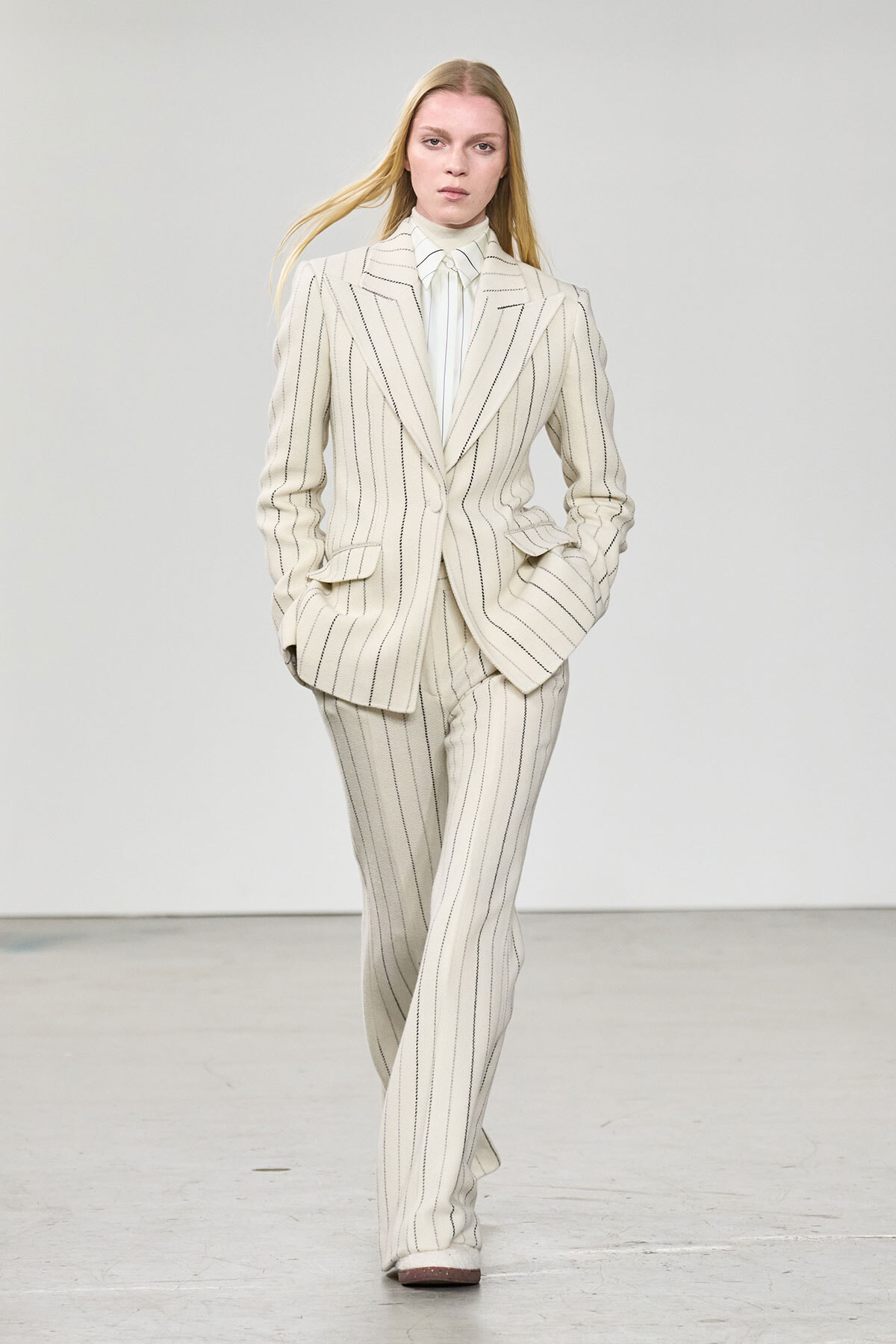 Model wearing a cream pinstripe suit (blazer and wide-leg trousers) walking on a runway.