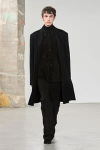 Male model walking a runway in an all-black ensemble: long coat over a black button-up shirt and dark pants.