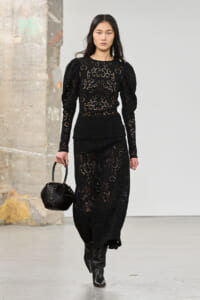 Model walking a runway in a black lace long-sleeve dress, pairing with black ankle boots and a woven black handbag.