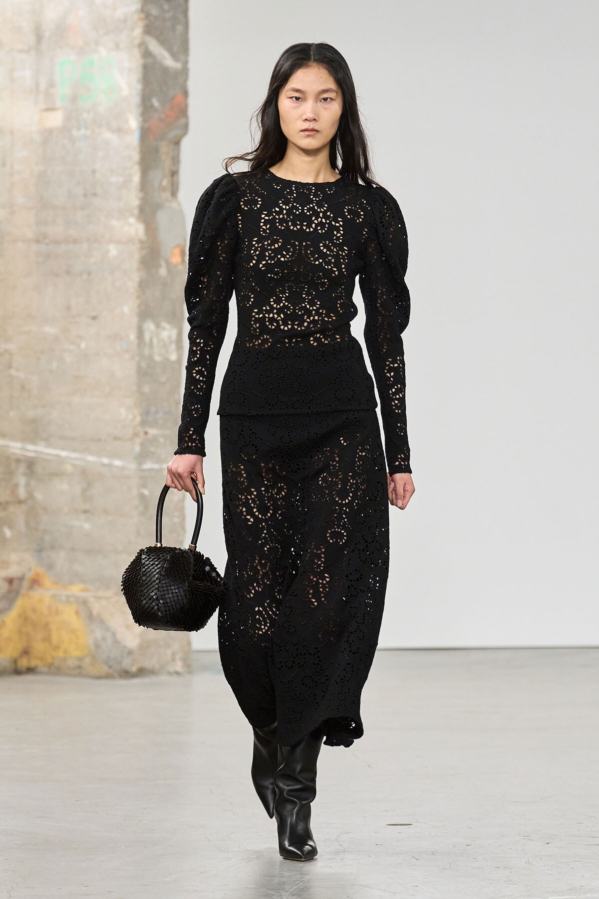 Model walking a runway in a black lace long-sleeve dress, pairing with black ankle boots and a woven black handbag.