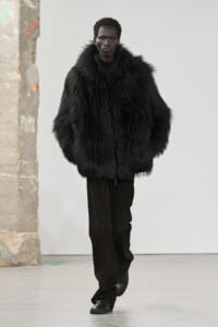 Model walking toward the camera in a large black fur coat and dark pants against a plain light background in a studio setting.