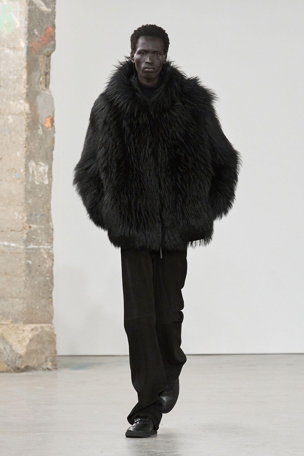 Model walking toward the camera in a large black fur coat and dark pants against a plain light background in a studio setting.
