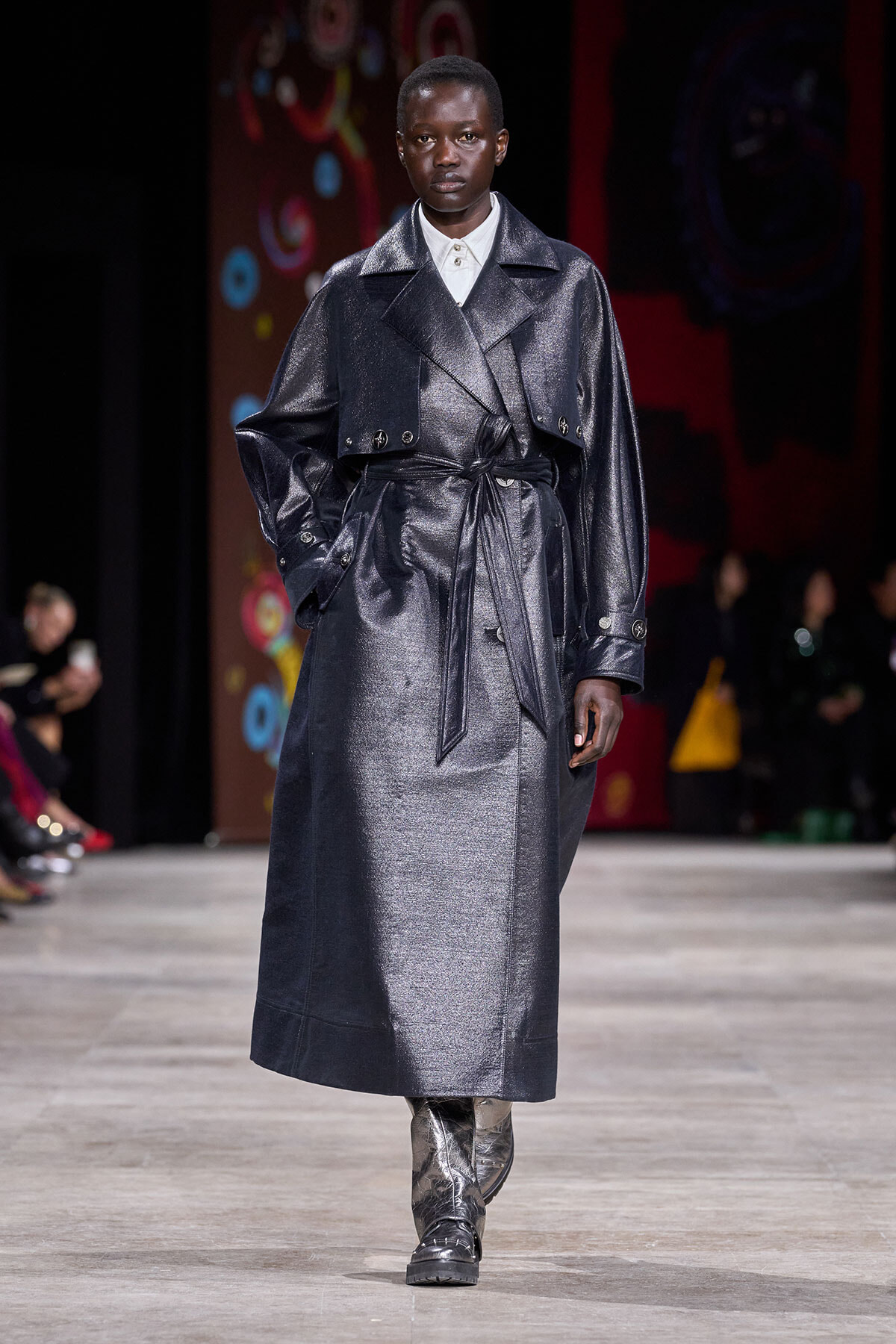 Model walks the runway in a shiny dark gray metallic trench coat cinched at the waist, white shirt peeking at the collar, and metallic boots.
