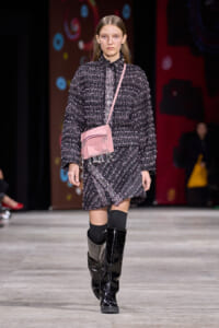 Model on a runway wearing a textured black-and-white tweed suit, knee-high black boots, and a pink crossbody bag.