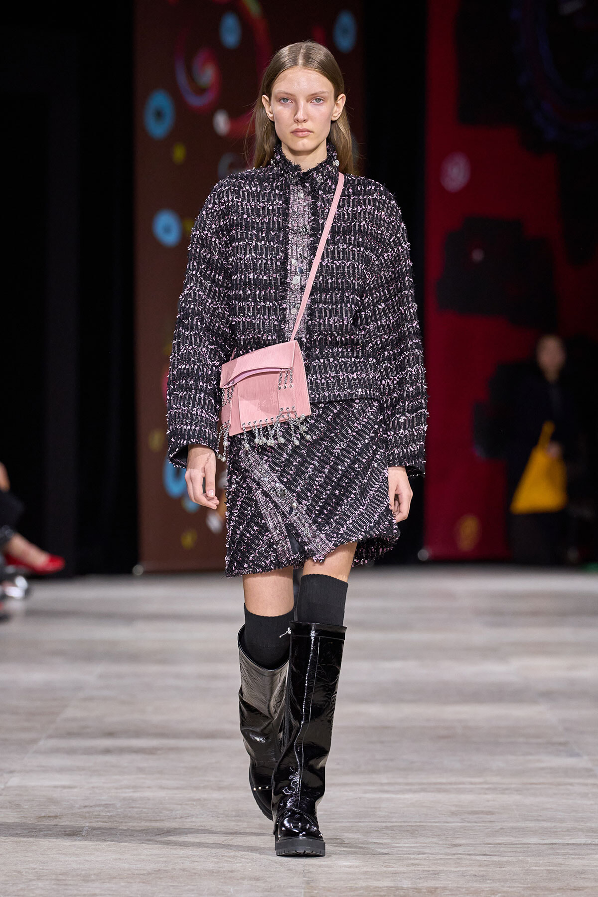 Model on a runway wearing a textured black-and-white tweed suit, knee-high black boots, and a pink crossbody bag.