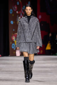 Model walking a runway in a metallic gray oversized coat and matching short skirt, black turtleneck, and knee-high patent boots, carrying a pink mini handbag