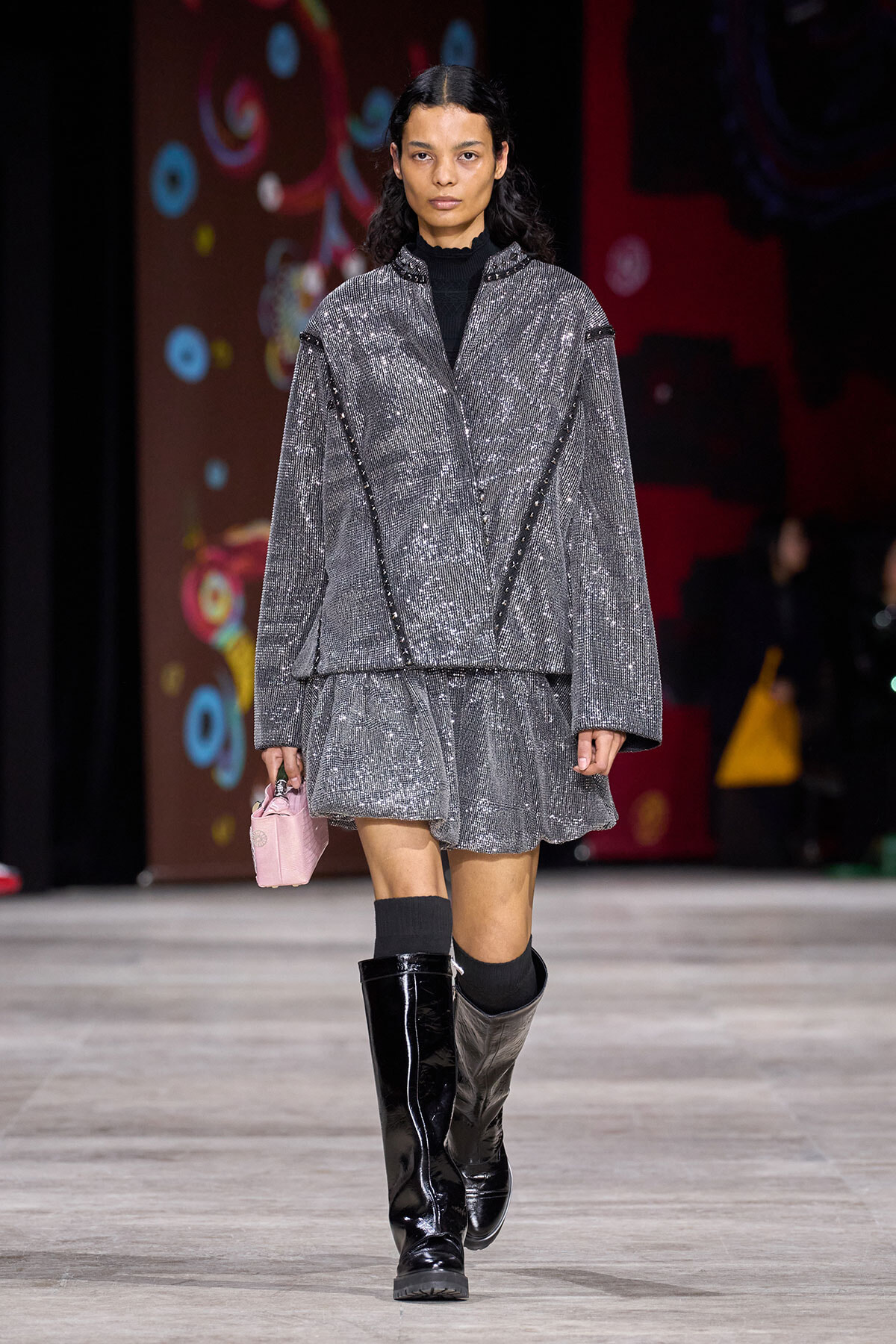 Model walking a runway in a metallic gray oversized coat and matching short skirt, black turtleneck, and knee-high patent boots, carrying a pink mini handbag