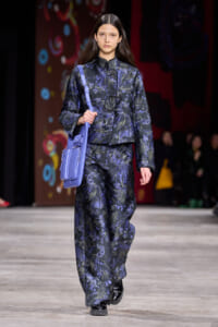 Model on a fashion show runway wearing a navy floral-pattern suit with a lavender crossbody bag slung over the shoulder.
