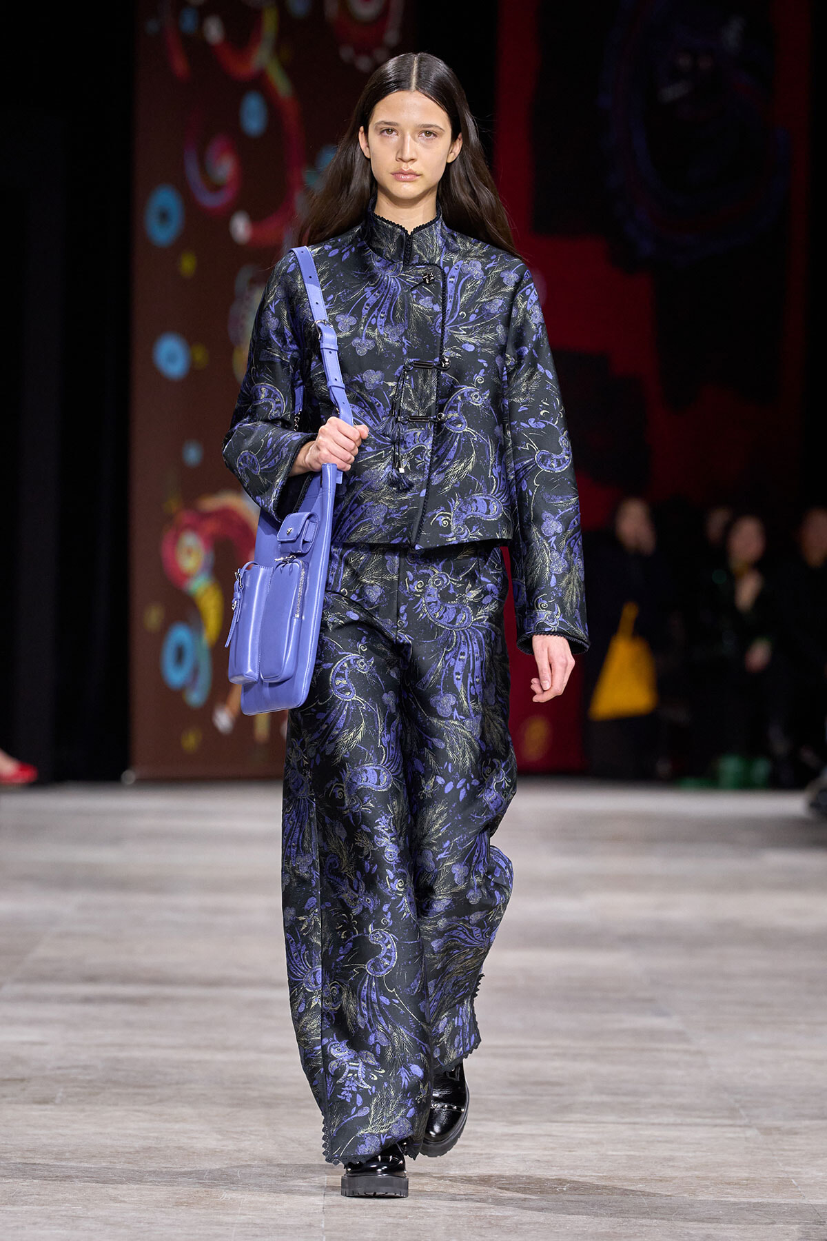 Model on a fashion show runway wearing a navy floral-pattern suit with a lavender crossbody bag slung over the shoulder.