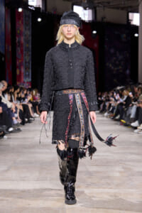 Model walking a fashion runway in a textured black coat, patterned belt, and knee-high black boots; wearing a black hat and fringe details visible at the hem.