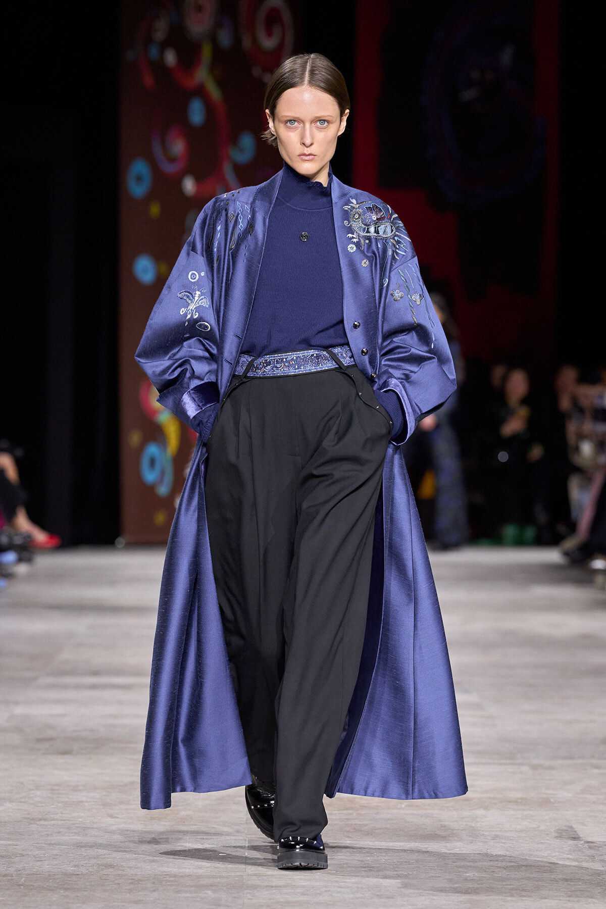 Model walks a runway wearing a blue satin robe-coat with embroidery over a dark blue turtleneck and black wide-leg pants.