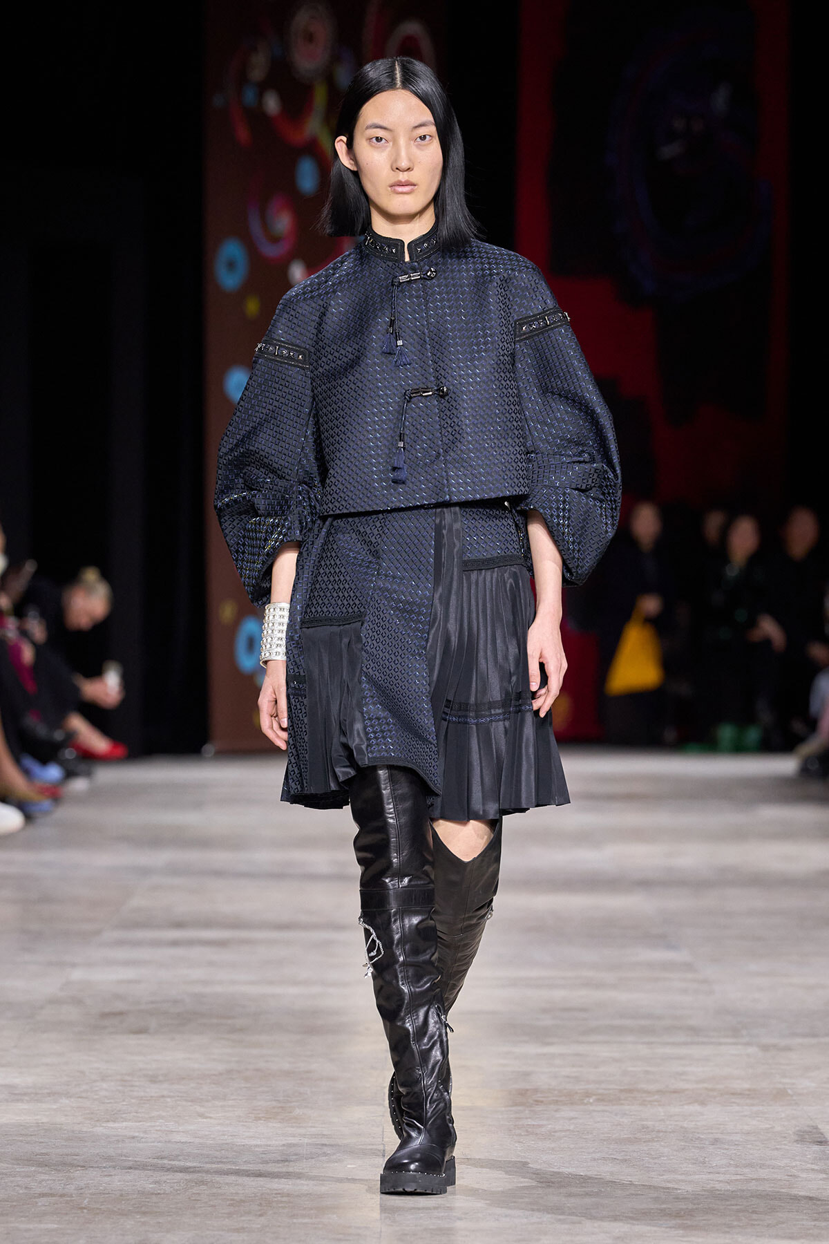 Model walking runway in navy textured top with puffed sleeves, matching pleated skirt, and over-the-knee black boots.
