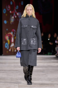 Model walks the runway in a charcoal gray coat with embroidered patches on chest and pockets, paired with knee-high black patent boots and a small blue handbag.