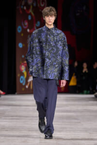 Male model on a runway wearing a dark blue patterned jacket and navy trousers, black shoes, mid-stride pose, backstage audience in background