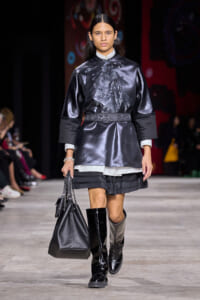 Model walks a runway in a metallic black tunic with floral embroidery, white cuffs peeking from sleeves, and a layered black skirt with knee-high boots, carrying a black handbag with chain straps.