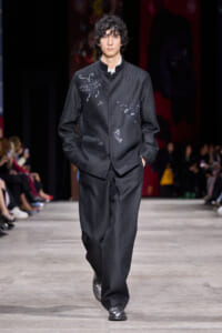 Male model walks a dark textured jacket and matching wide trousers down a fashion runway, subtle embroidered designs on the jacket.
