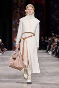 Runway model in an ivory layered coat with a high-neck sweater, a tan belt, and a large beige handbag.