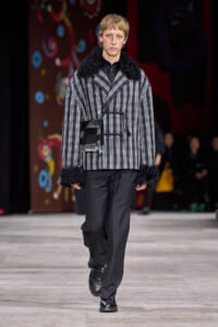 Male model walking runway in a gray plaid coat with black faux fur cuffs and a black crossbody bag worn at the waist.