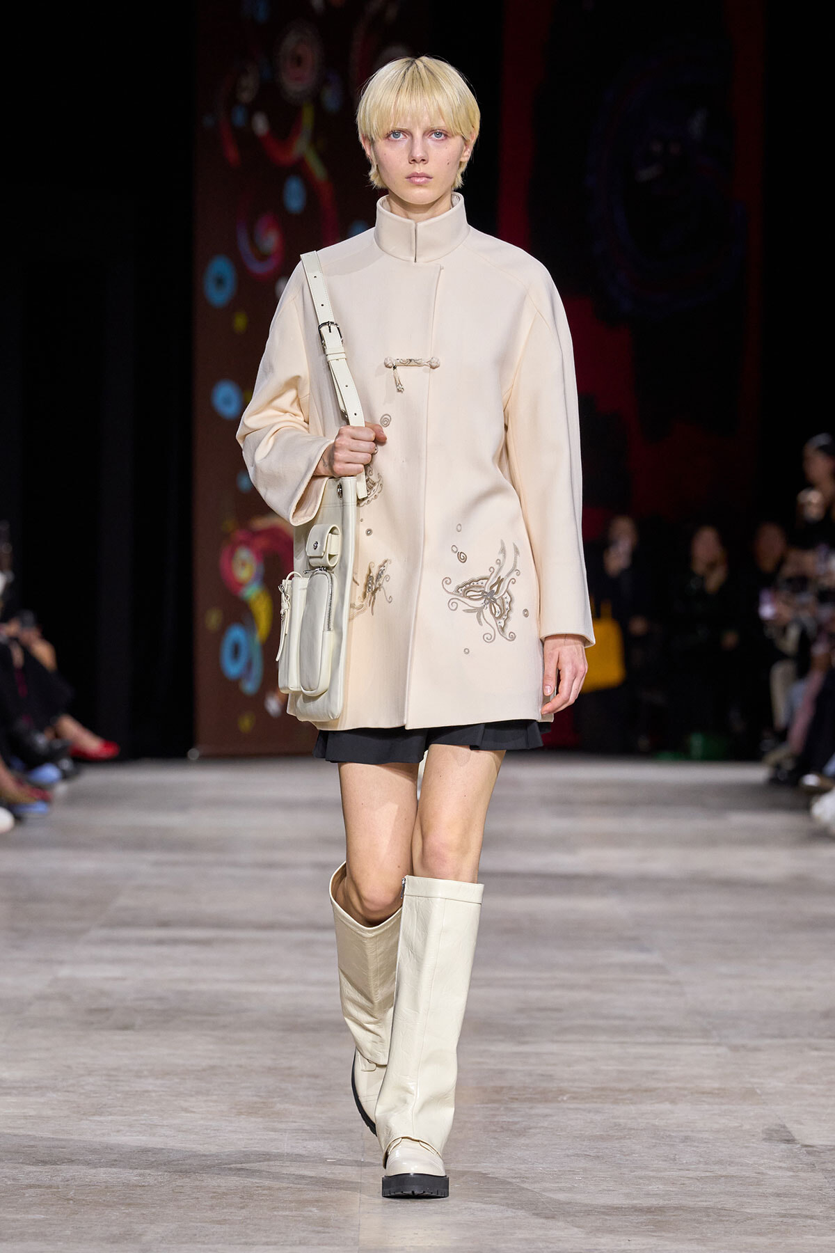 Model on a runway wearing a cream high-neck coat with subtle embroidery, carrying a white crossbody bag and wearing knee-high cream boots.