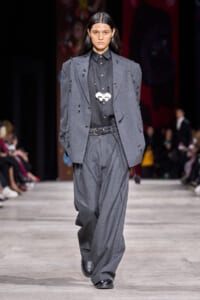 Model walking a gray oversized suit on a runway, wearing a black shirt and a chunky necklace, with hands in pockets.