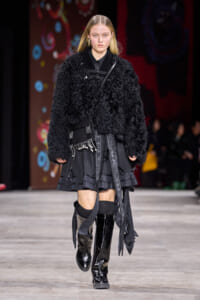 Female model walks a runway in a voluminous black boucle coat, layered dark skirt, and shiny knee‑high boots.