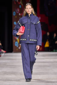 Female model walks a fashion show runway in a dark blue denim jacket and wide-leg jeans, holding a red clutch.