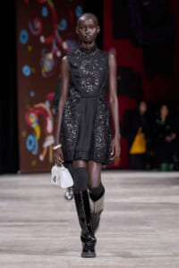 Model walking a fashion runway in a black sequined dress with a high neck and decorative embroidery, paired with tall black boots and a small white handbag.