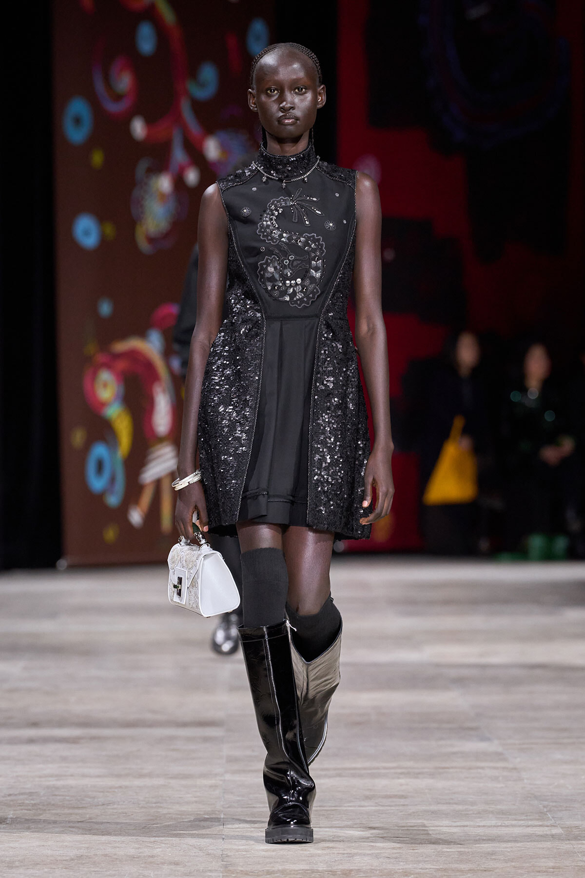 Model walking a fashion runway in a black sequined dress with a high neck and decorative embroidery, paired with tall black boots and a small white handbag.