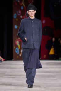 Male model walking a fashion runway in a dark blue oversized coat with matching pants and a flowing pleated skirt-like layer, black loafers, and a black beret-style hat.