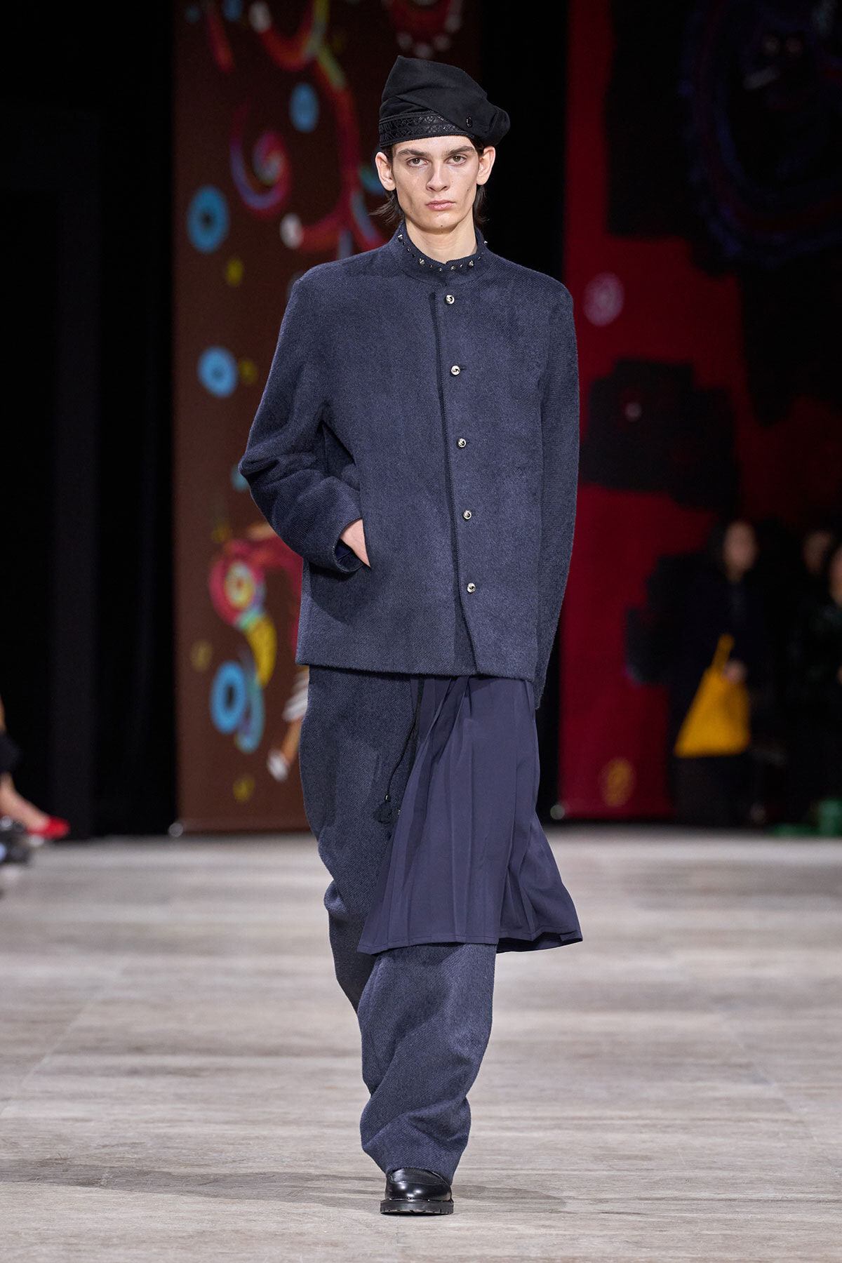Male model walking a fashion runway in a dark blue oversized coat with matching pants and a flowing pleated skirt-like layer, black loafers, and a black beret-style hat.
