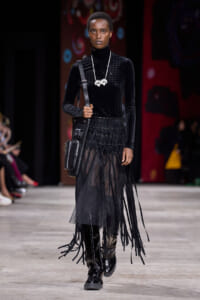 Model walking the runway in an all-black ensemble: velvet turtleneck, sheer fringe skirt, knee-high boots, and a black shoulder bag with a white statement necklace.