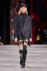 Model walking the runway in a black sequined coat with fringe at the hem, paired with a black turtleneck, leather cap, and knee-high patent boots.