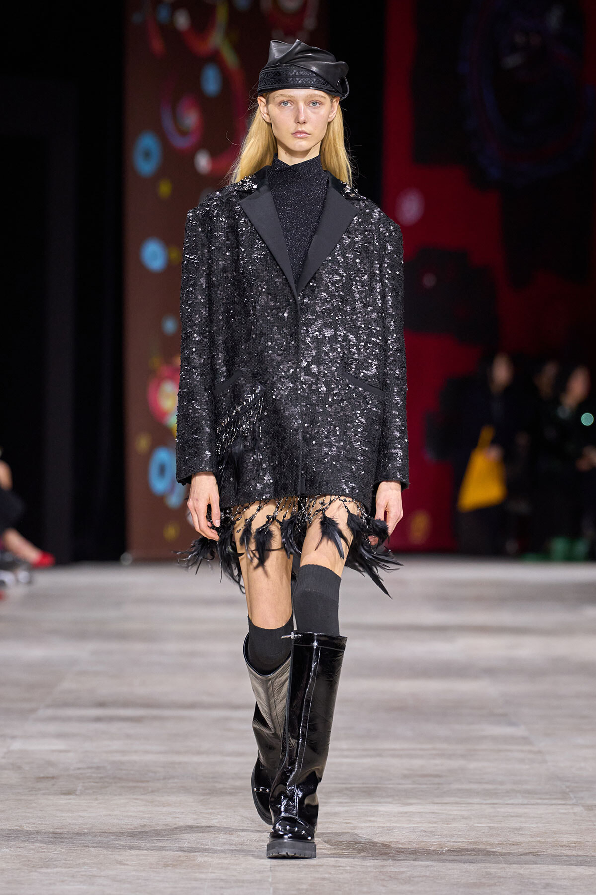 Model walking the runway in a black sequined coat with fringe at the hem, paired with a black turtleneck, leather cap, and knee-high patent boots.
