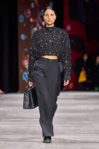 Model walking a runway in a black beaded cropped jacket and high-waisted wide-leg trousers, carrying a black handbag.