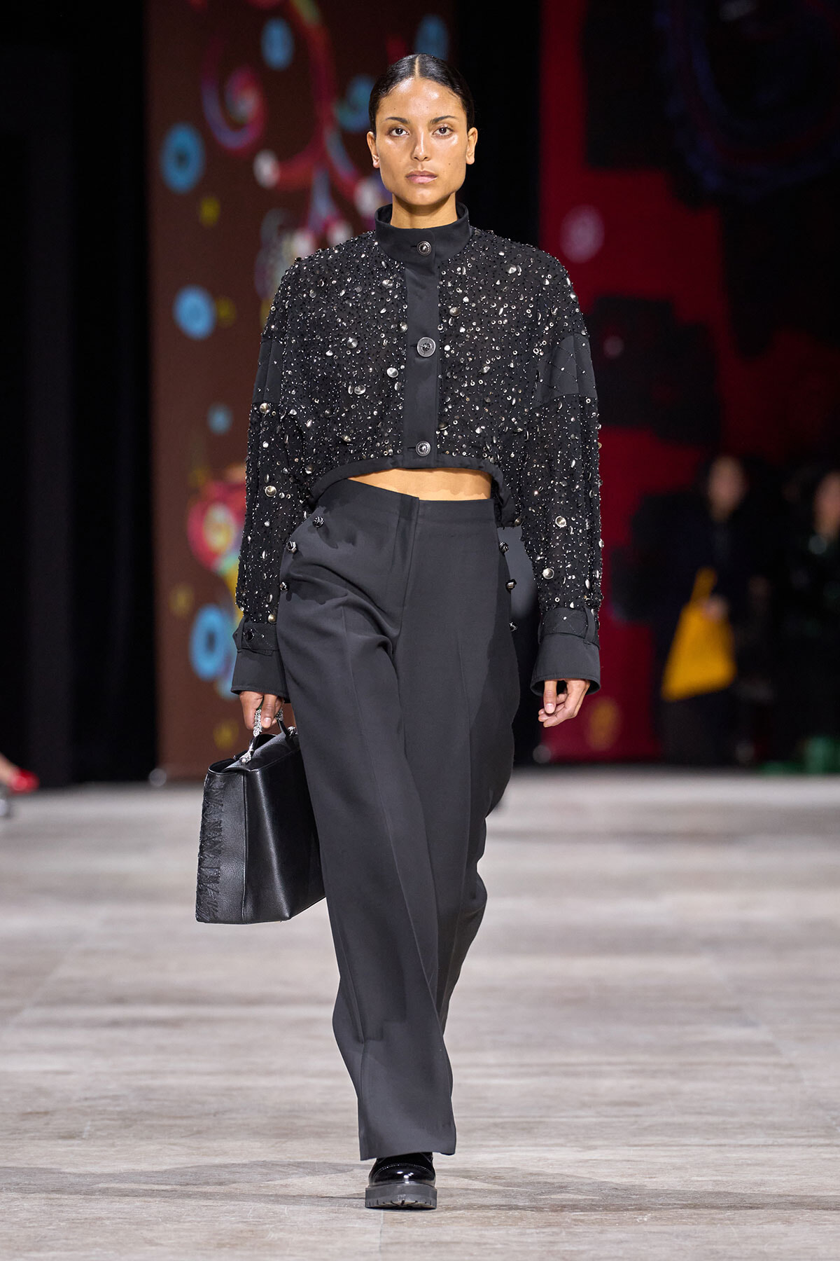 Model walking a runway in a black beaded cropped jacket and high-waisted wide-leg trousers, carrying a black handbag.