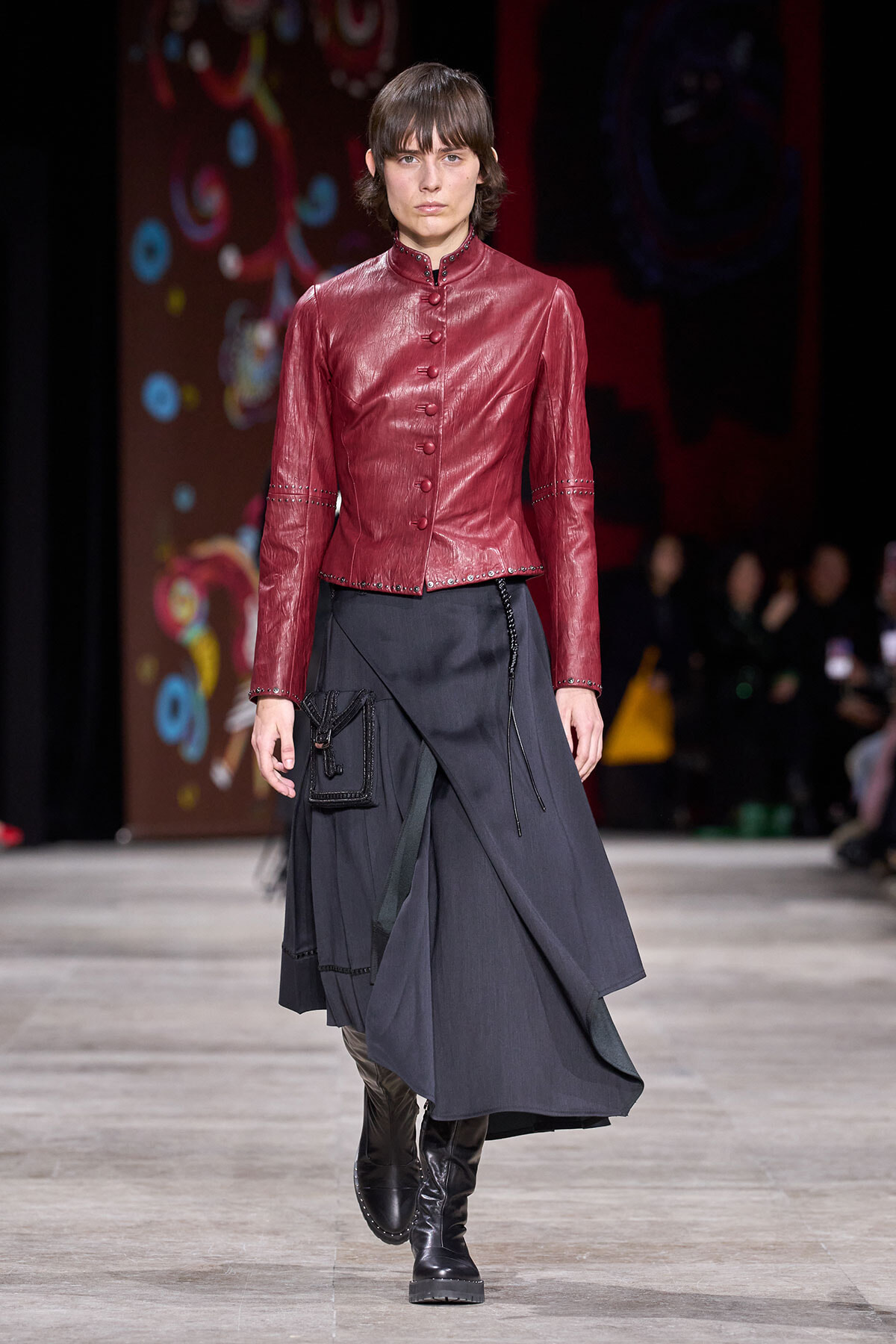 Model walking the runway in a red leather button-front jacket and a dark asymmetrical wrap skirt with a small black pouch and knee-high boots.