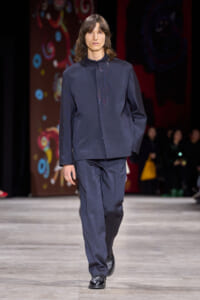 Male model walks a fashion runway in a navy tailored jacket and matching trousers with subtle front embroidery on the jacket screen-printed in dark threads.