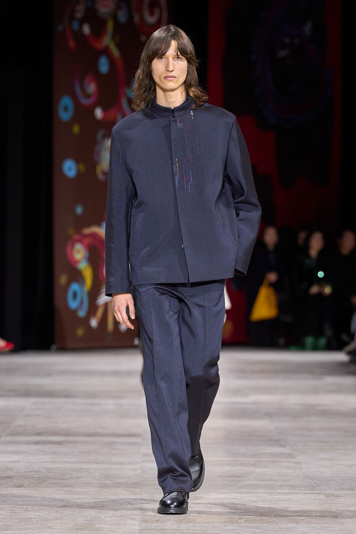 Male model walks a fashion runway in a navy tailored jacket and matching trousers with subtle front embroidery on the jacket screen-printed in dark threads.