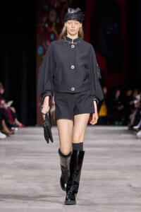 Model walks the runway in a dark gray oversized jacket with puff sleeves, matching shorts, black knee-high patent boots, and a black cap with pearl necklace.